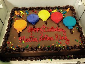 Square shaped cake with chocolate frosting, five balloons and the words "Happy Birthday Martin Luther King"
