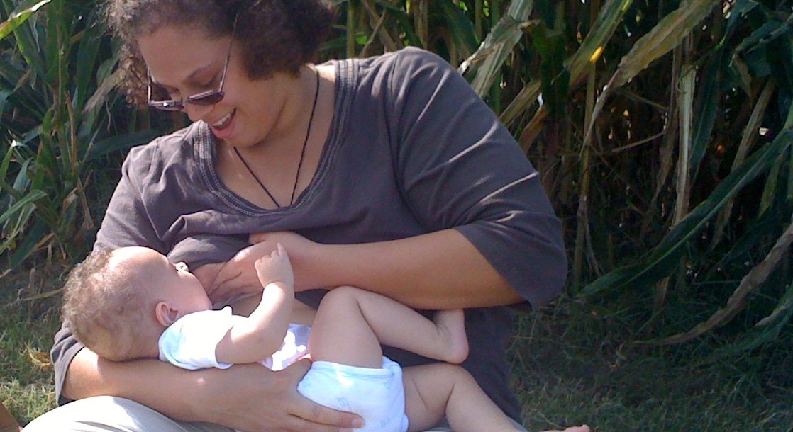 Woman sitting cross-legged on the ground, smiling while breastfeeding and looking at her infant.